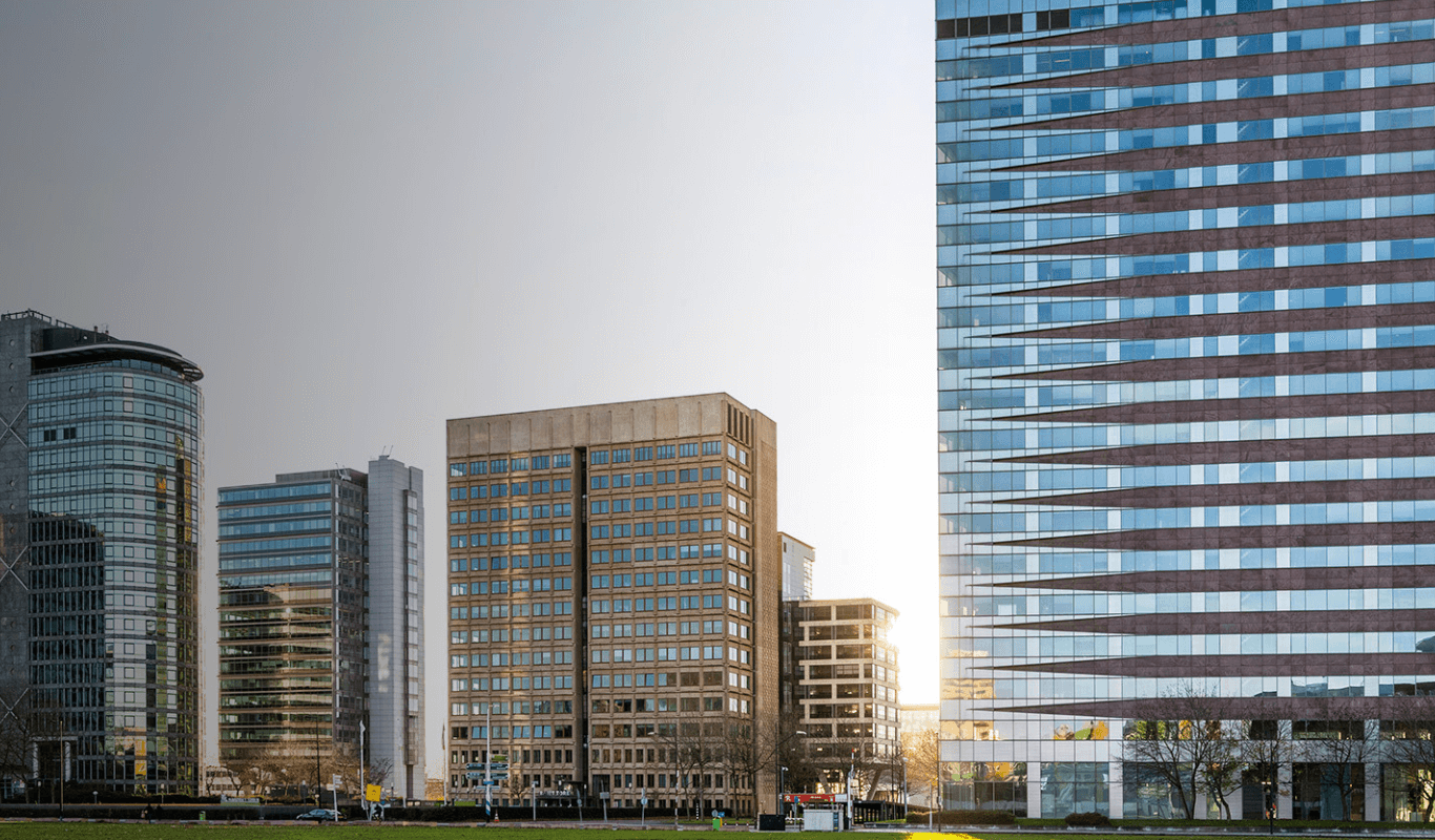 Foto van het B-Vital kantoorpand in Amsterdam Zuidoost. Het moderne gebouw met roodbruine en glazen horizontale banen staat centraal, omringd door andere hoge kantoorgebouwen. De zon schijnt er net achter langs.