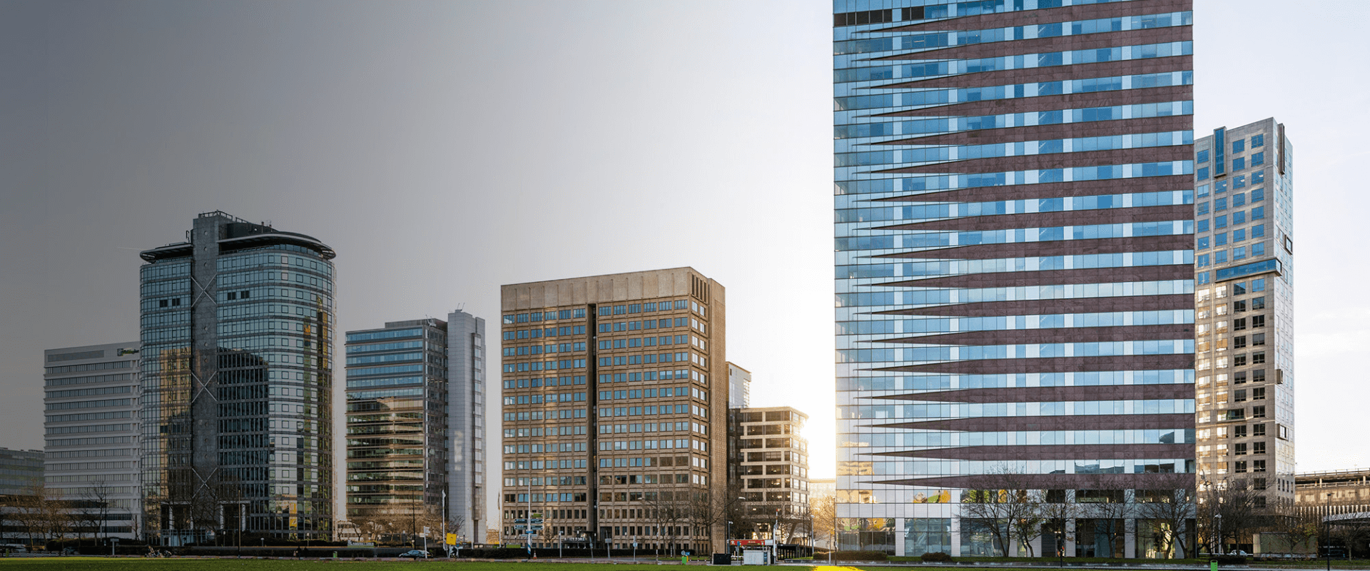 Foto van het B-Vital kantoorpand in Amsterdam Zuidoost. Het moderne gebouw met roodbruine en glazen horizontale banen staat centraal, omringd door andere hoge kantoorgebouwen. De zon schijnt er net achter langs.