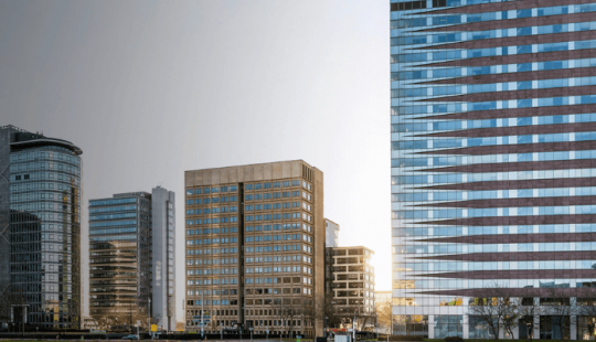 Foto van het B Vital kantoorpand in Amsterdam Zuidoost. Het moderne gebouw met roodbruine en glazen horizontale banen staat centraal, omringd door andere hoge kantoorgebouwen. De zon schijnt er net achter langs.