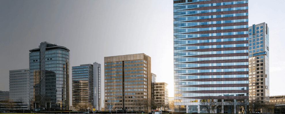 Foto van het B Vital kantoorpand in Amsterdam Zuidoost. Het moderne gebouw met roodbruine en glazen horizontale banen staat centraal, omringd door andere hoge kantoorgebouwen. De zon schijnt er net achter langs.