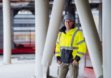 Man met veiligheidshelm en felgele reflecterende jas staat op een bouwlocatie tussen betonnen pilaren. Hij kijkt in de camera en draagt werkkleding.