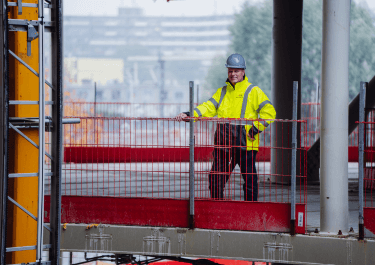 Man met veiligheidshelm en fluorescerend gele jas staat op een bouwplaats achter een rood hekwerk. Op de achtergrond zijn bouwmaterialen en vage contouren van gebouwen zichtbaar.