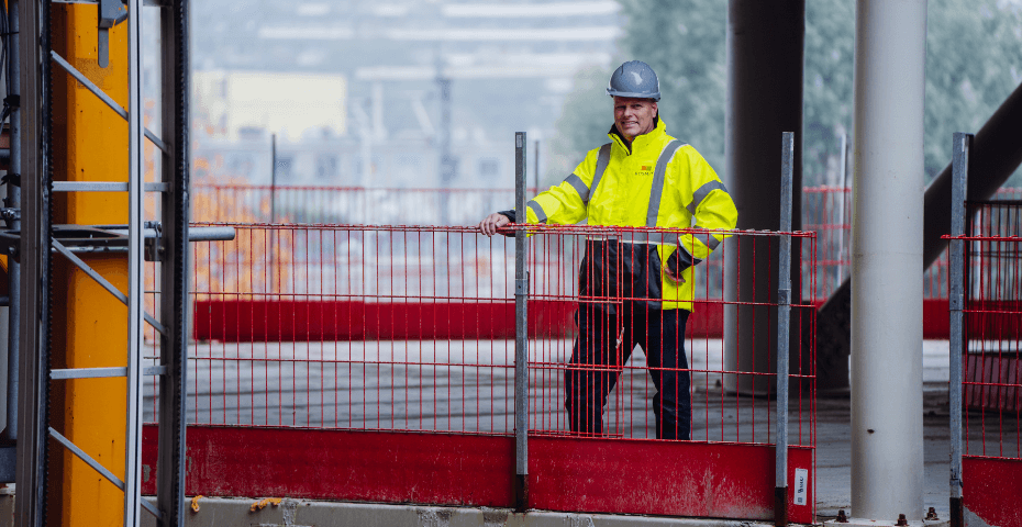 Man met veiligheidshelm en fluorescerend gele jas staat op een bouwplaats achter een rood hekwerk. Op de achtergrond zijn bouwmaterialen en vage contouren van gebouwen zichtbaar.