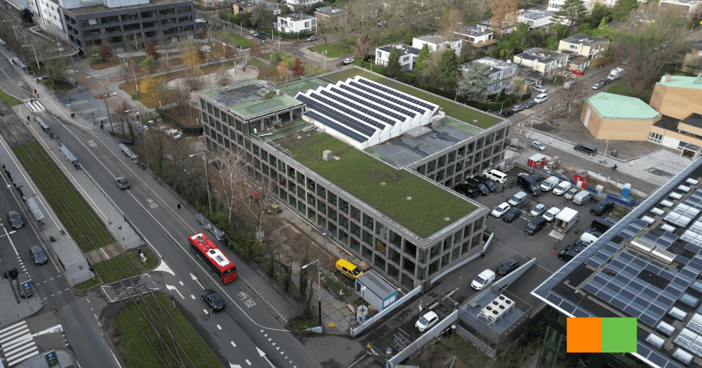 Luchtfoto van het Strawinskyhuis in Amsterdam, een duurzaam kantoorgebouw met een groen dak en zonnepanelen, gelegen naast een drukke weg en trambaan.
