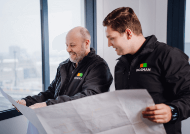 Twee medewerkers van Bosman Bedrijven bekijken samen bouwtekeningen in een lichte kantooromgeving.