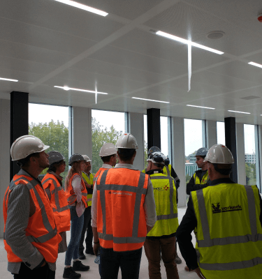 Groep medewerkers van verschillende bedrijven met veiligheidshelmen en hesjes tijdens een projectbespreking op een bouwlocatie.