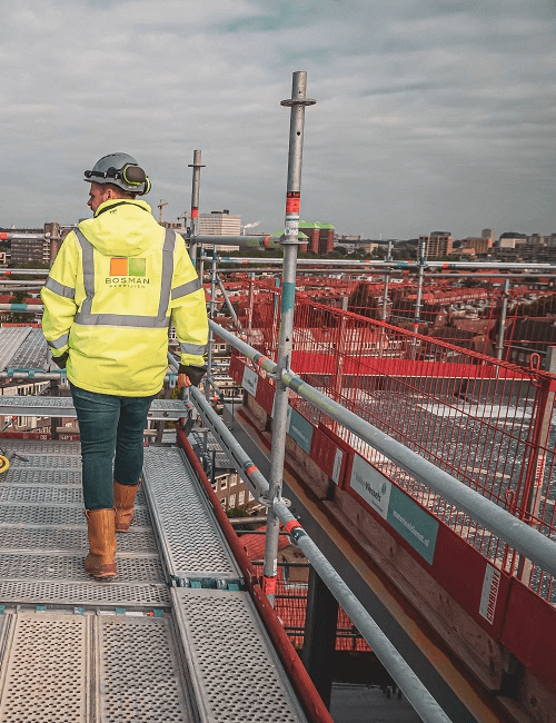 Een Bosman-medewerker loopt op een bouwplaats op hoogte, met zicht op een stedelijke omgeving en draagt veiligheidskleding.