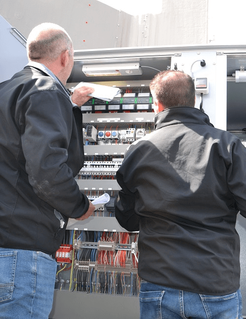 Twee medewerkers controleren een open elektrische schakelkast met bedrading en componenten tijdens een technische inspectie.