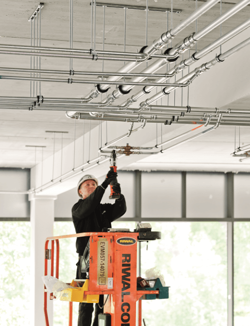 Een man werkt op een hoogwerker aan plafondinstallaties, omringd door complexe leidingstructuren.