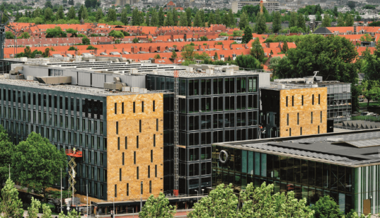 Buitenaanzicht van EDGE Stadium in Amsterdam, een modern en duurzaam kantoorgebouw met glazen gevels en gele accenten, omringd door groen.