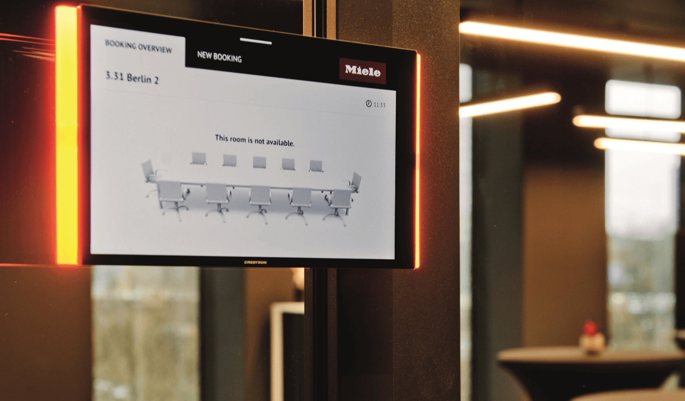 Digitaal reserveringsscherm voor een vergaderruimte bij Miele X in Amsterdam, gemonteerd op een zwarte pilaar in een modern kantoorinterieur.