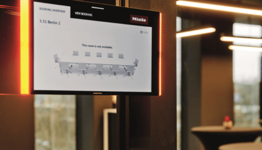 Digitaal reserveringsscherm voor een vergaderruimte bij Miele X in Amsterdam, gemonteerd op een zwarte pilaar in een modern kantoorinterieur.