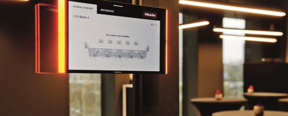 Digitaal reserveringsscherm voor een vergaderruimte bij Miele X in Amsterdam, gemonteerd op een zwarte pilaar in een modern kantoorinterieur.