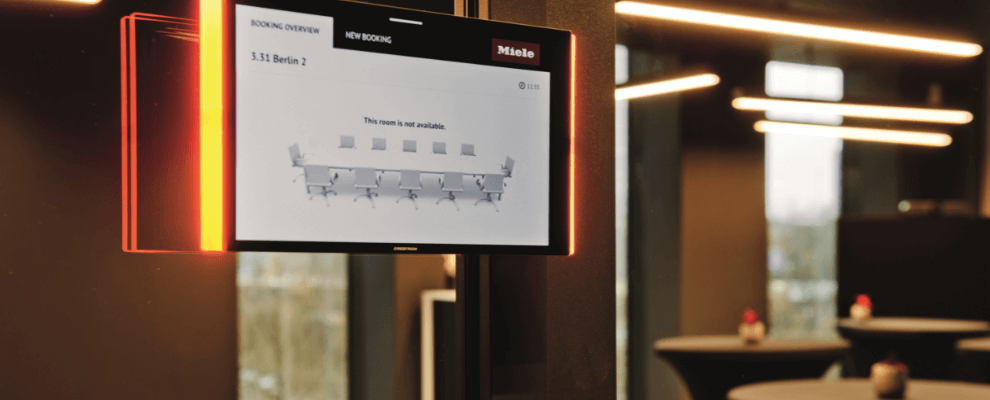 Digitaal reserveringsscherm voor een vergaderruimte bij Miele X in Amsterdam, gemonteerd op een zwarte pilaar in een modern kantoorinterieur.