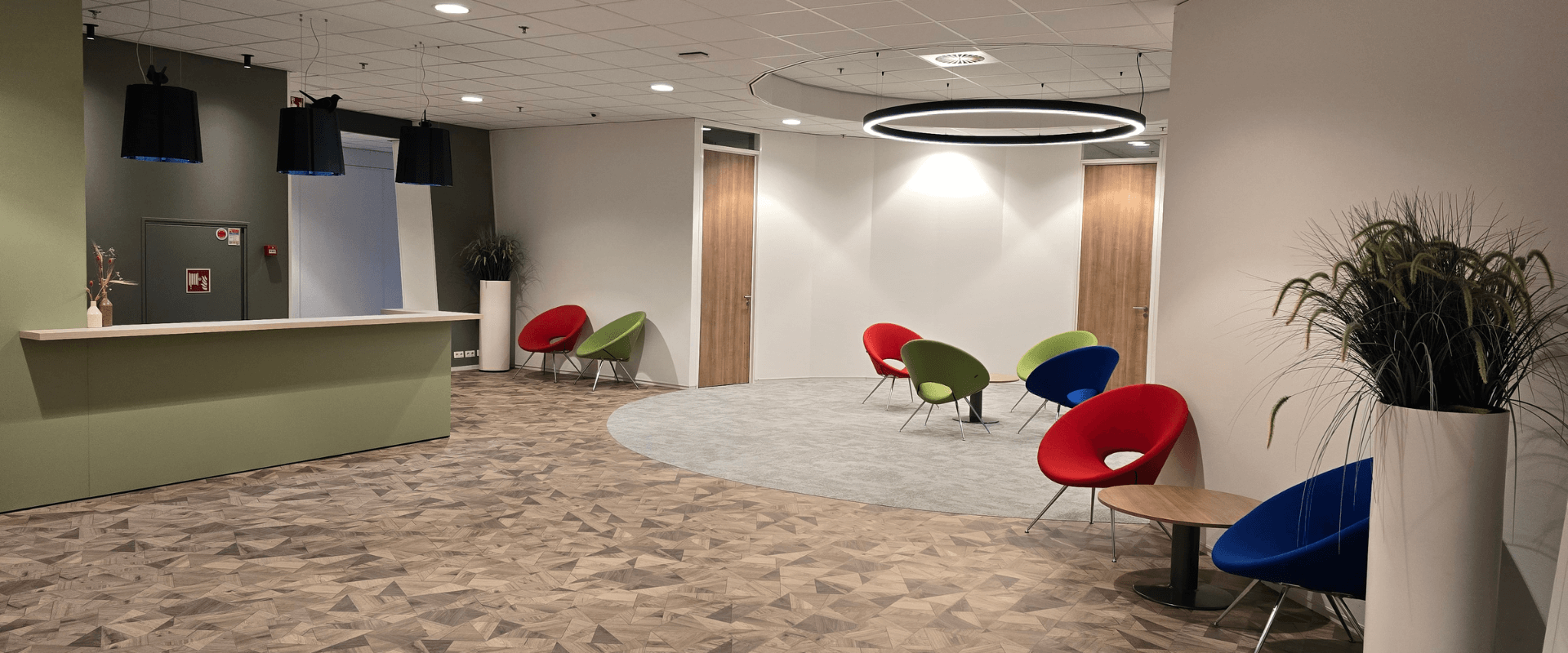 Moderne en sfeervolle ontvangstruimte met een receptiebalie, kleurrijke designstoelen en warme houten vloer, ontworpen voor een uitnodigende en comfortabele werkomgeving.