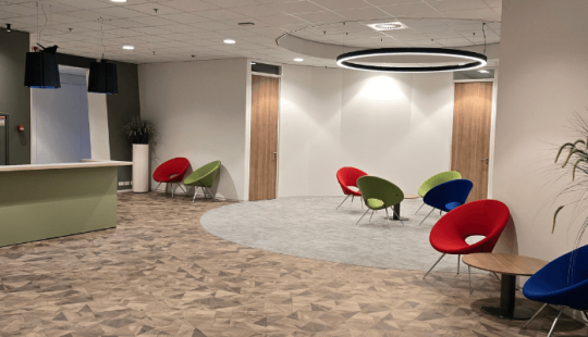 Moderne en sfeervolle ontvangstruimte met een receptiebalie, kleurrijke designstoelen en warme houten vloer, ontworpen voor een uitnodigende en comfortabele werkomgeving.