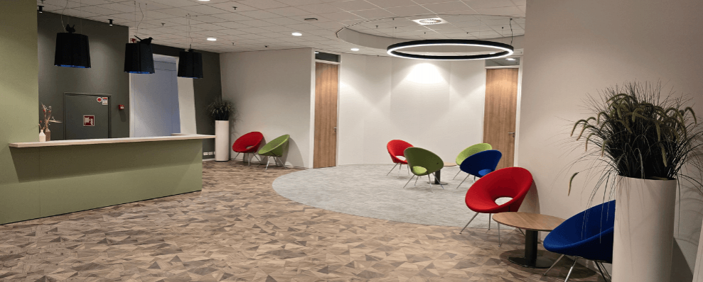Moderne en sfeervolle ontvangstruimte met een receptiebalie, kleurrijke designstoelen en warme houten vloer, ontworpen voor een uitnodigende en comfortabele werkomgeving.