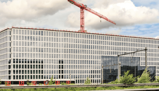 Bouw van Tripolis Park in Amsterdam, een modern en duurzaam kantoorgebouw met glazen gevel en rode bouwkraan op de achtergrond.