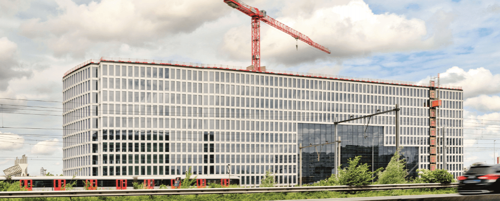 Bouw van Tripolis Park in Amsterdam, een modern en duurzaam kantoorgebouw met glazen gevel en rode bouwkraan op de achtergrond.