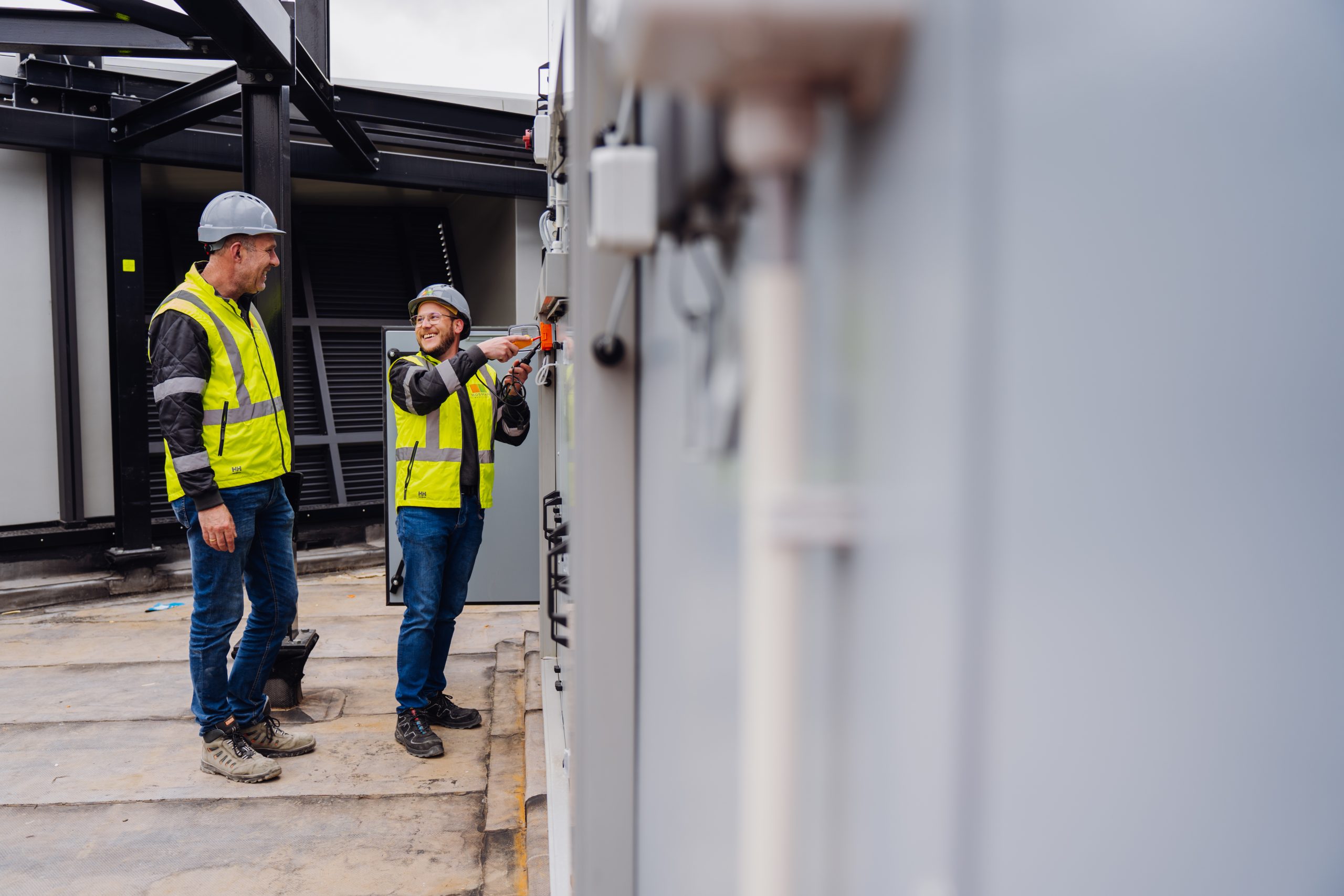 Twee mannen in veiligheidskleding en helmen werken lachend aan een elektrisch paneel op een industriële locatie. Eén man bedient het paneel met een schroevendraaier terwijl de ander toekijkt. Beiden dragen gele veiligheidshesjes en staan op een betonnen vloer met metalen constructies op de achtergrond.