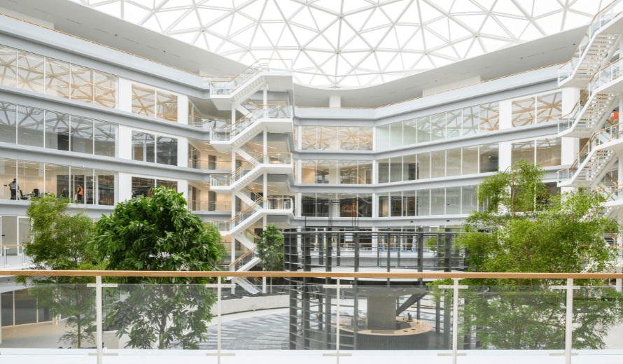 Een ruime, moderne kantoorlobby met meerdere verdiepingen, veel glas en natuurlijk licht. In het midden staat een ronde structuur, omringd door jonge bomen en brede looproutes.