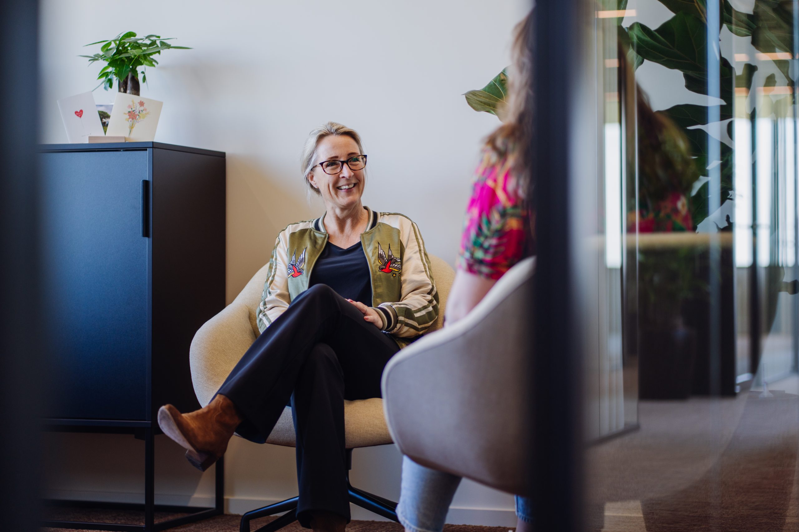 Een vrouw zit glimlachend in een fauteuil tijdens een gesprek in een modern kantoor, deels gezien door een glazen wand.