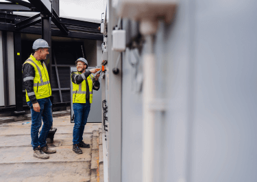 Twee mannen in veiligheidskleding en helmen werken lachend aan een elektrisch paneel op een industriële locatie. Eén man bedient het paneel met een schroevendraaier terwijl de ander toekijkt. Beiden dragen gele veiligheidshesjes en staan op een betonnen vloer met metalen constructies op de achtergrond.