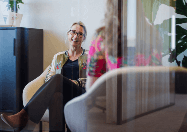 Een vrouw zit glimlachend in een fauteuil tijdens een gesprek in een modern kantoor, deels gezien door een glazen wand.