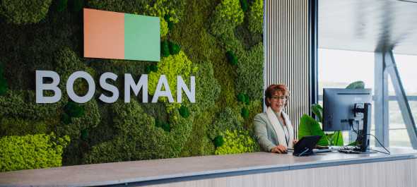 Receptioniste bij de balie van Bosman, met moswand en logo op de achtergrond, klaar om bezoekers te ontvangen.