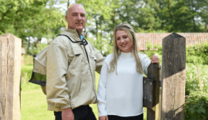 Imker Eddy en dochter Quinty poseren samen bij een houten hek op een groene, landelijke locatie.