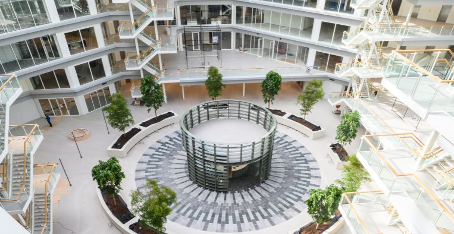 Een ruime, moderne kantoorlobby met meerdere verdiepingen, veel glas en natuurlijk licht. In het midden staat een ronde structuur, omringd door jonge bomen en brede looproutes.