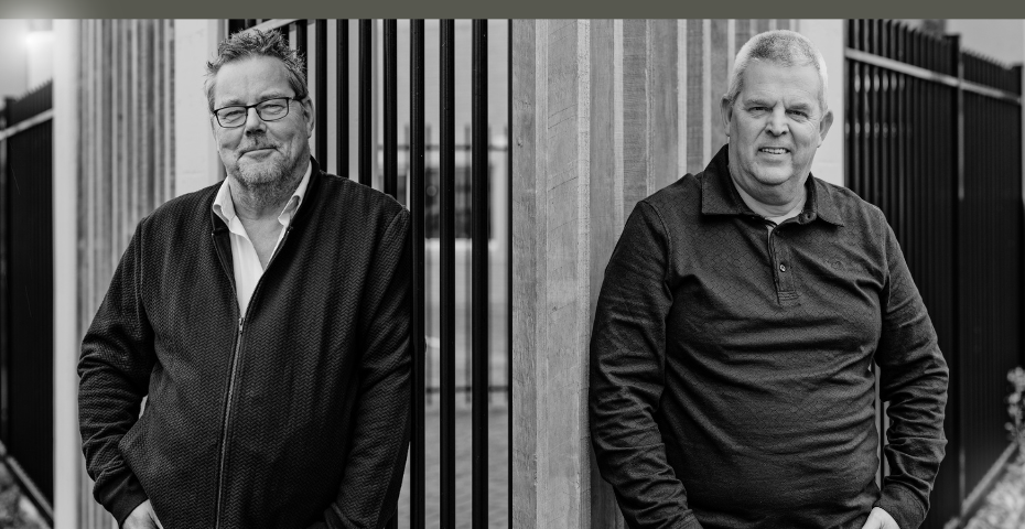 Twee mannen leunen ontspannen tegen een houten paal en metalen hekwerk, gekleed in donkere kleding, kijkend naar de camera in een zwart-wit portret.