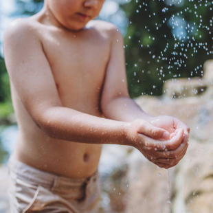 Kind houdt spelenderwijs water vast onder een fontein of waterval, met de focus op de handen en waterdruppels.