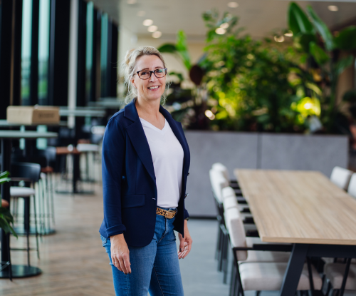Medewerkster, HR Business Partner, staat glimlachend in een moderne kantoorruimte naast een vergadertafel.
